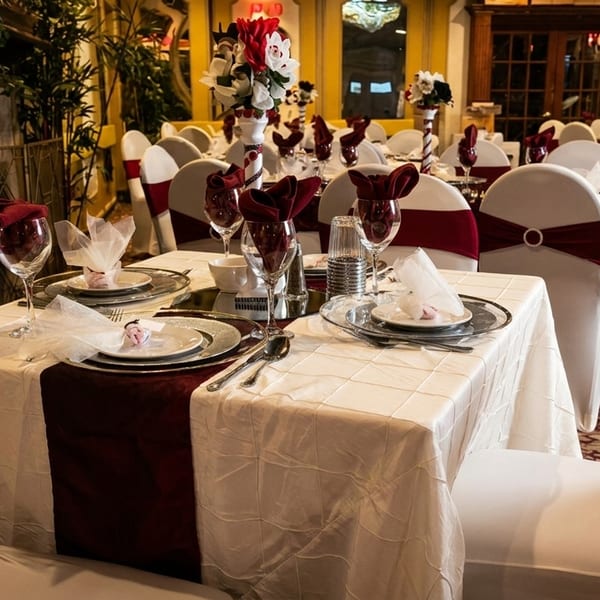 Elegantly set dining tables with white tablecloths, burgundy runners, and matching chair sashes in a restaurant.