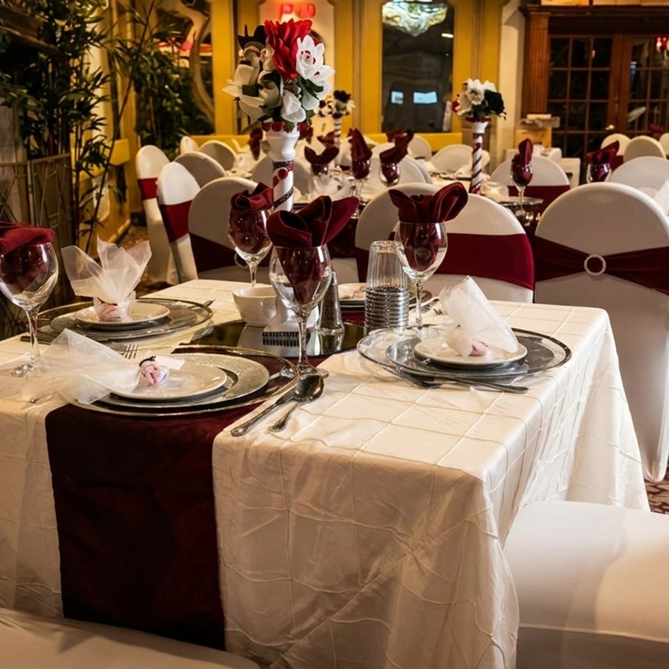 Elegantly set dining tables with white tablecloths, burgundy runners, and matching chair sashes in a restaurant.
