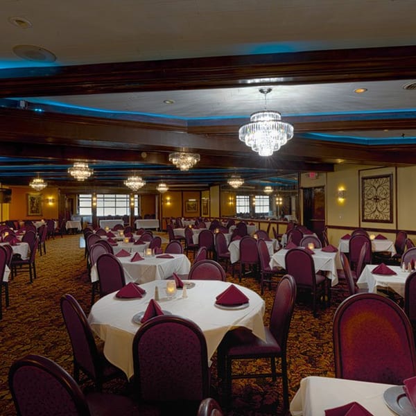 Elegant dining room with white-clothed tables, maroon chairs, chandeliers, and blue cove lighting.
