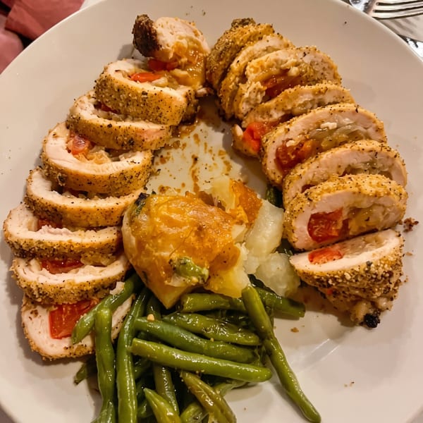 Sliced chicken roulade with red peppers and onions, green beans, and a baked potato on a white plate.
