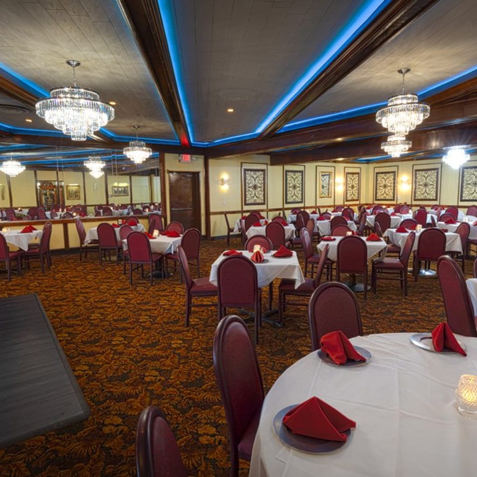 Spacious restaurant dining room with many tables set with white tablecloths, red napkins, and chandeliers.