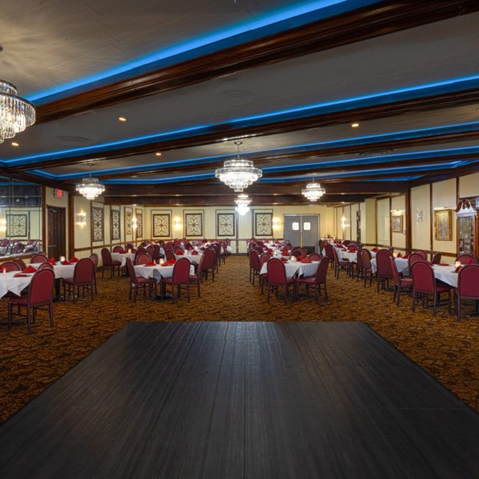 A large banquet hall with many tables set with white tablecloths and red chairs, under blue accent lighting.